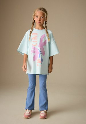 Junges Mädchen mit geflochtenem Haar, trägt ein übergroßes pastellblaues T-Shirt mit Vogelprint, blaue ausgestellte Hosen und pinke Turnschuhe, steht neutral.