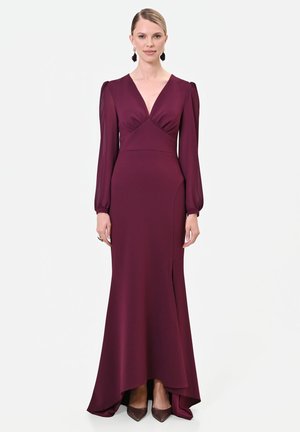 Donna che indossa un abito lungo fino a terra, a maniche lunghe, di un profondo color bordeaux, con scollo a V e una leggera apertura frontale, abbinato a scarpe scure con punta appuntita.