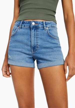 Woman wearing blue denim rolled-hem shorts and an olive green sleeveless ribbed top, shown from mid-torso to mid-thigh.
