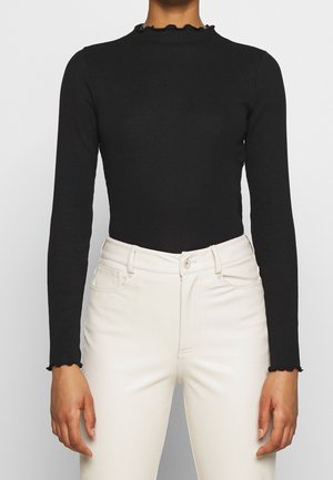 Person iført en tætsiddende sort langærmet top med flæsekrave og flæsemanchetter, sammen med højtaljede hvide jeans, stående mod en ensfarvet baggrund.