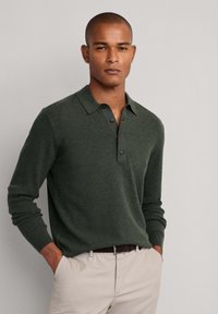 Tamnozelena polo košulja dugih rukava s opuštenim krojem, okovratnikom na dugmad i rebrastom teksturom, uparen s svijetlosmešavim hlačama i smešim remenom.