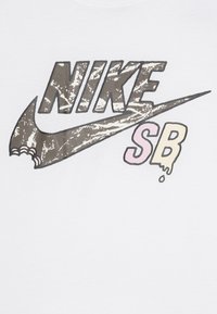 Balta T-kreklu ar grafisku apdruku, kurā redzams "NIKE" izsistā brūnā un melnā tonī, un apakšā ir "SB" pasteļrozā un dzeltenā krāsā, izdrukāta centrāli.