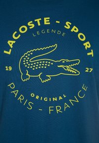 Logo crocodile Lacoste jaune avec le texte "Lacoste Sport," "Legende," "Original," et "Paris France 1927" sur un fond en tissu bleu.