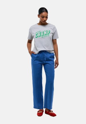 T-shirt en coton gris avec un texte vert indiquant "DEVIENS LE CHAMPION QUE TU ES DÉJÀ", assorti à un pantalon en lin bleu à jambes larges et des ballerines rouges.