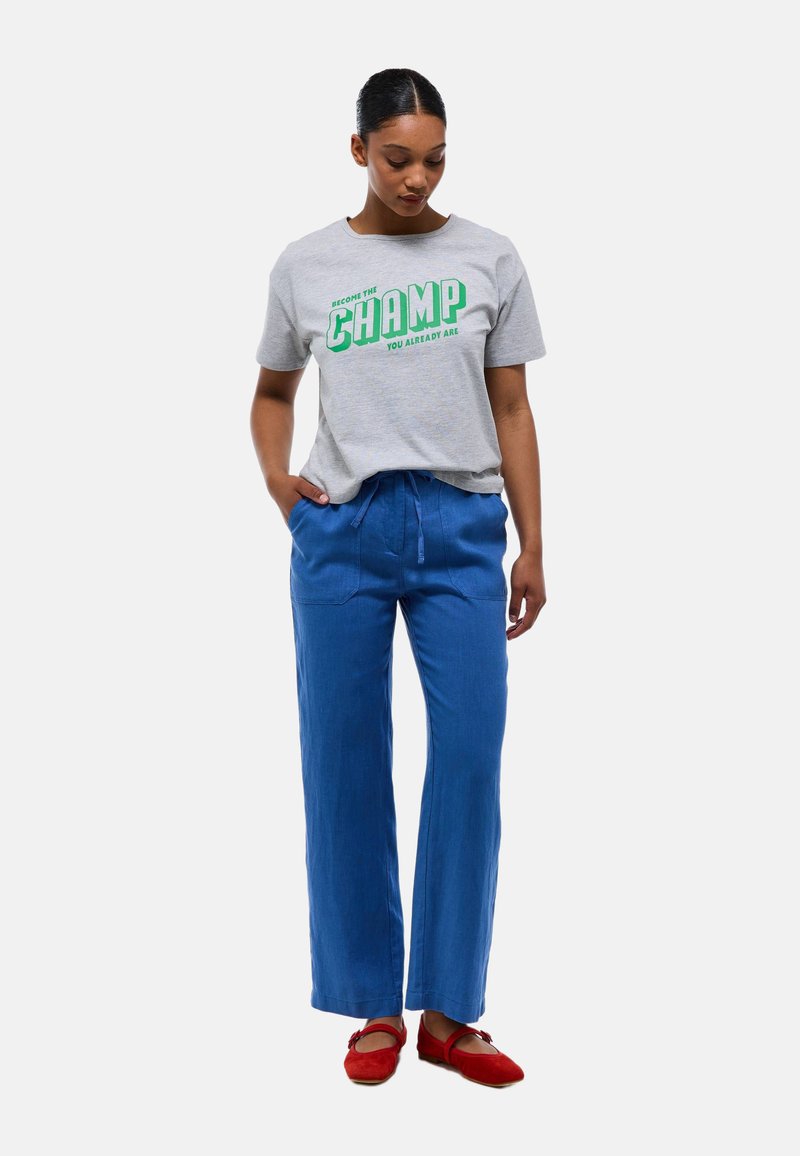 T-shirt en coton gris avec un texte vert indiquant "DEVIENS LE CHAMPION QUE TU ES DÉJÀ", assorti à un pantalon en lin bleu à jambes larges et des ballerines rouges.