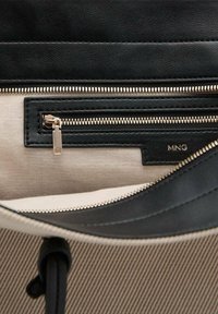 Intérieur d'un sac noir et beige avec plusieurs poches zippées, présentant un design en cuir lisse et en tissu texturé. Accents en métal doré visibles.