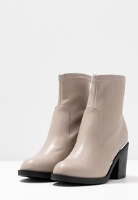 Botas de tobillo beige con puntera redondeada, tacones gruesos negros y costuras visibles, sobre un fondo blanco liso.
