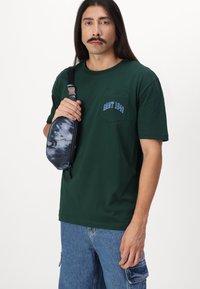 Mørkegrøn bomulds t-shirt med en brystlomme og blå broderet tekst "GANT 1948". Holder en tie-dye mønstret sling bag med lynlås.