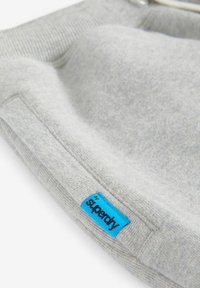 Šedá mikina z látky s jemnou textúrou. Obsahuje modrú nálepku s textom "Superdry". Hladký golier a manžety. Minimalistický dizajn.