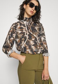 Blusa de mangas longas com um padrão floral escuro e terroso, gola plissada, e punhos arregaçados, combinada com calças verde-azeitona.