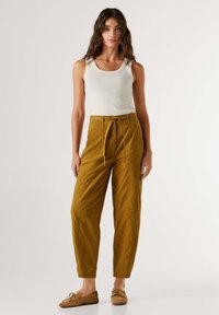 Frau steht vor einfachem Hintergrund, trägt ein weißes ärmelloses Oberteil, senfgelbe Hosen mit Kordelzug und braune Mokassins.