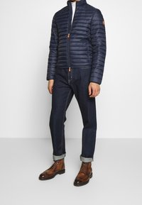 Marineblå pufferjakke med quiltet design, opretstående krave og kontrastfarvet orange lynlåstræk, kombineret med mørkeblå denimjeans og brune støvler.