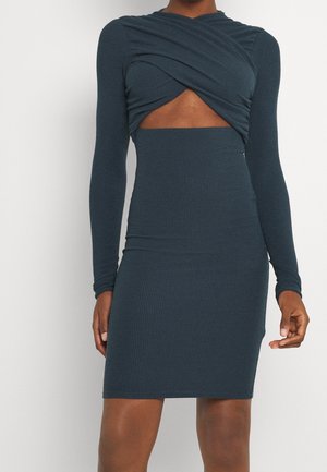 Donker teal ribgebreide jurk met lange mouwen en een gedraaid uitsnijdingsdetail ter hoogte van de taille, halflang aan een staande persoon.