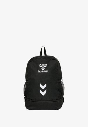 Sac à dos noir avec un extérieur en tissu lisse, arborant un logo Hummel blanc et un design en chevrons, ainsi que des poches latérales en mesh et une poignée sur le dessus.