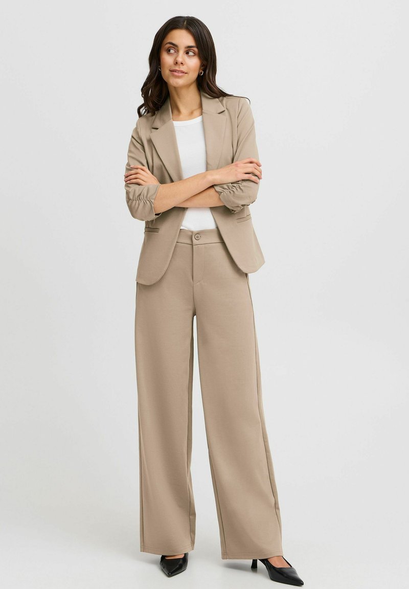 Beige skräddarsydd blazer med uppvikta ärmar, i kombination med vida byxor. Slät textur, enkel knappstängning och tidlös design. Svarta klackar.