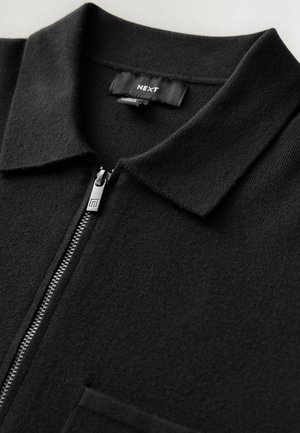Schwarzes Strick-Polohemd mit Reißverschluss vorne, spitzem Kragen und Brusttasche. Hat eine glatte Textur und minimales Design.