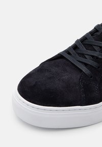 Baskets en daim bleu marine avec des lacets plats gris foncé, une semelle en caoutchouc blanche et des détails de couture subtils sur la surface supérieure.