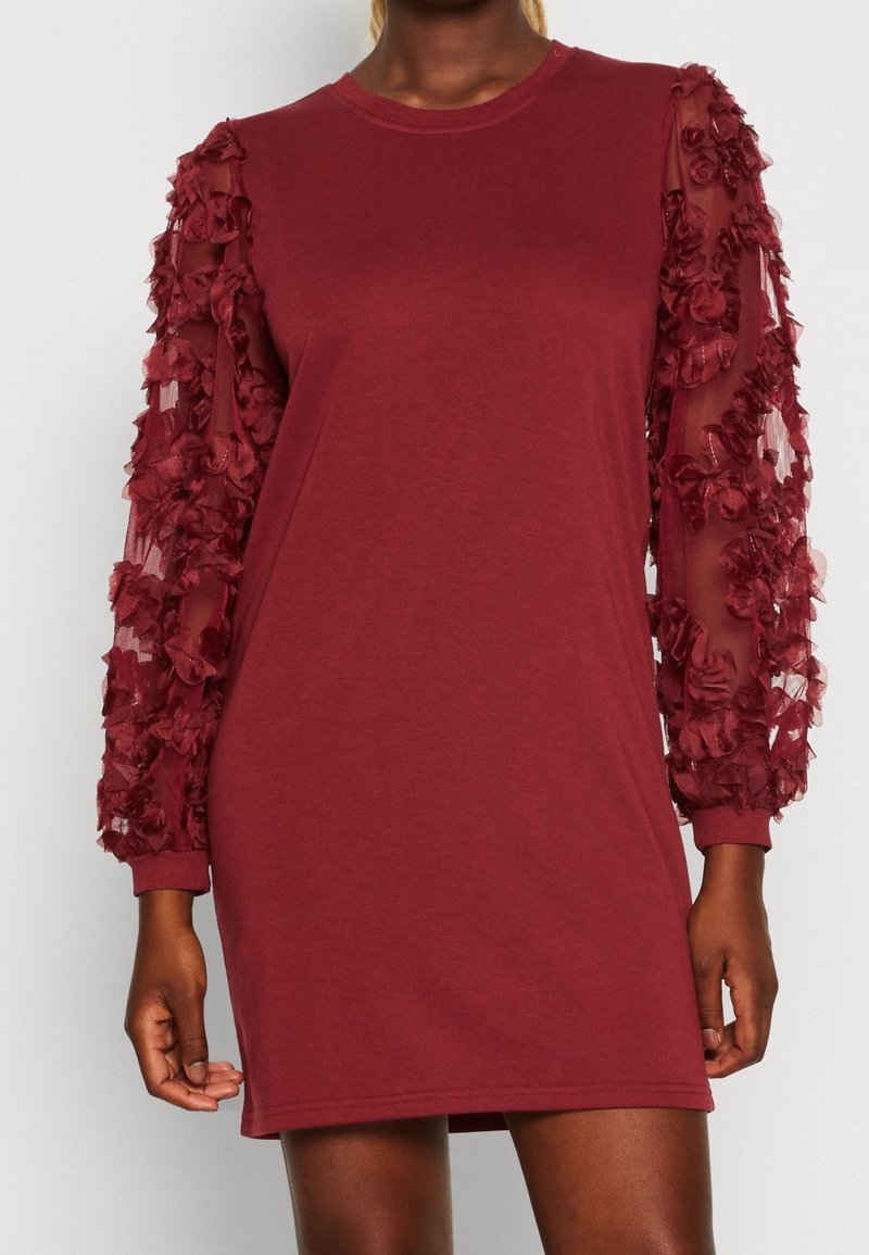 Femme portant une robe bordeaux avec des manches longues transparentes décorées d'appliques florales, debout devant un fond uni.