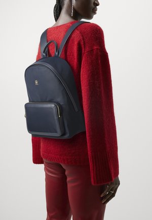 Zaino in nylon blu navy con forma arrotondata, spallacci imbottiti e tasca frontale con zip. Presenta un dettaglio del logo in oro e una texture liscia.