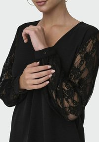Czarna bluzka z dekoltem w kształcie litery V, z długimi koronkowymi rękawami. Rękawy mają kwiatowy wzór koronki oraz zapięcie na guzik. Gładka faktura materiału.