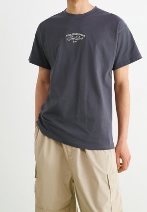 Mann trägt ein dunkelgraues Nike Sportswear T-Shirt mit dem Text "Just Do It" und beige Cargoshorts, steht mit einer Hand in der Tasche.