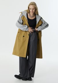 Gelber Trenchcoat mit grauem Hoodie, ausgestattet mit großen Knöpfen und Taschen. Kombiniert mit weit geschnittenen, dunkelgrauen Hosen und schwarzen Sneakern.