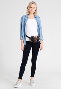 Fanny pack de camuflaje negro y marrón con cierre de cremallera, llevado sobre una camisa blanca y combinado con pantalones oscuros ajustados y zapatillas blancas.