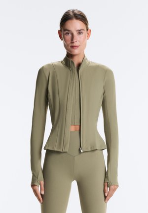 Žena nosiaca priliehavú olivovo zelenú bundu so zipsom a zodpovedajúce legíny, stojaca pred jednoliatym bielym pozadím, čelom vpred.