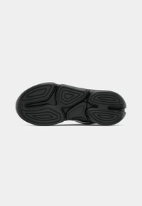 Schwarze Sneaker-Sohle mit geometrischen Profilmustern, von unten vor einem schlichten weißen Hintergrund fotografiert.