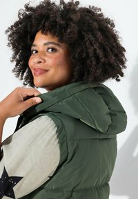 Gilet imbottito verde con cappuccio, caratterizzato da una texture liscia e un design trapuntato. Indossato sopra una camicia chiara con motivo a stelle.