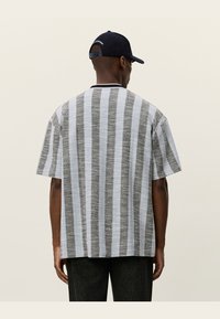 Camisa de mangas curtas masculina com riscas verticais cinzas e pretas, textura em trama, decote redondo e corte descontraído. Boné preto usado.