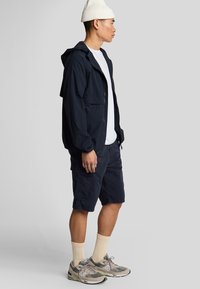 Marineblauwe lichtgewicht jas met capuchon, gecombineerd met bijpassende shorts. Witte T-shirt eronder, beige geribde beanie en grijze sneakers met accenten.
