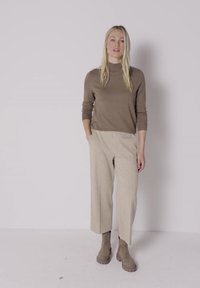 Femme aux longs cheveux blonds porte un haut marron à manches longues, un pantalon large beige et des bottines taupe, debout devant un fond clair uni.