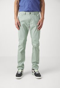 Hombre que lleva pantalones rectos de color verde claro, zapatillas negras y blancas, y una camisa azul, de pie contra un fondo blanco liso.