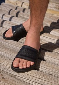 Sandali neri in gomma con due larghe fasce decorate con motivi geometrici, poggiati su una superficie in legno con un arco plantare visibile.