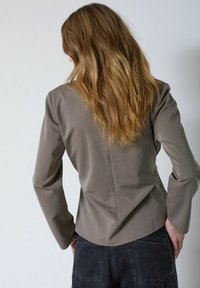 Mujer con cabello largo y ondulado, vistiendo un blazer gris ajustado y vaqueros oscuros, de pie con las manos en las caderas, frente a una pared gris claro.
