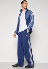 Trenirka tamnoplave boje s jaknom na patent s naglascima u svijetloplavoj boji i bijelim prugama, uparena s odgovarajućim hlačama i crnim cipelama.