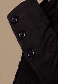 Close-up van een zwarte suède jas aan de mouw met drie grote zwarte knopen en zichtbare stiksels op een neutrale achtergrond.