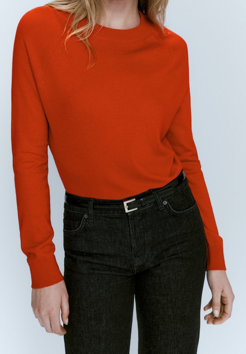 Femme portant un pull ajusté à manches longues orange vif rentré dans un jean noir taille haute avec une ceinture noire.