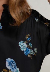 Blouse noire à fleurs avec des roses bleues et grises, texture soyeuse, manches courtes et coupe décontractée avec des détails plissés aux épaules.