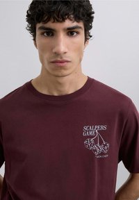 Camiseta de algodón burdeos con cuello redondo que presenta un gráfico de dados y el texto "SCALPERS GAME" en blanco en el área del pecho izquierdo.