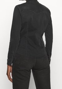 Persona che indossa una camicia in denim nera aderente e jeans neri, vista da dietro, su uno sfondo bianco semplice.