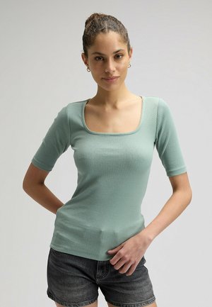 Femme aux cheveux bouclés attachés en chignon, portant des boucles d'oreilles créoles dorées, un haut côtelé vert clair à encolure carrée et un short en denim noir, posant.