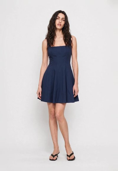 Femme aux cheveux foncés ondulés portant une robe bleu marine sans manches et des sandales noires à talons, debout devant un fond gris clair uni.
