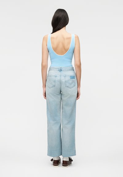 Γυναίκα με μακριά σκούρα μαλλιά, γυρισμένη πίσω, φοράει ανοιχτό μπλε κοντό τοπ με ανοιχτή πλάτη, ανοιχτό μπλε τζιν παντελόνι με φαρδιά μπατζάκια και καφέ παπούτσια.