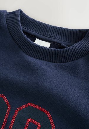 Tengerészkék pulóver bordázott kerek nyakkal, nagy piros hímzett szám dizájnnal. Puha tapintású, egyszínű kivitelben.