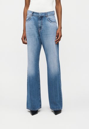 Persoon draagt lichtblauwe wijde jeans en zwarte puntige schoenen, staande tegen een effen witte achtergrond.