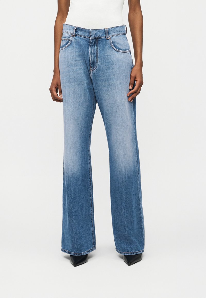 Άτομο που φοράει ανοιχτό μπλε τζιν με φαρδιά πόδια και μαύρα μυτερά παπούτσια, όρθιο μπροστά σε απλό λευκό φόντο.