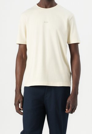 Mann, der ein cremefarbenes Kurzarm-T-Shirt mit kleinem "BOSS"-Logo und dunkelblaue Hose trägt, vor einem schlichten weißen Hintergrund steht.