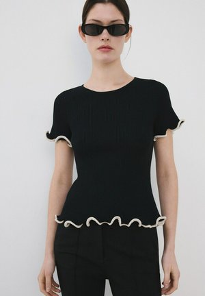 Joven mujer vestida con camiseta negra acanalada con bordes blancos con volantes y pantalones negros, posando frente a un fondo claro y liso.